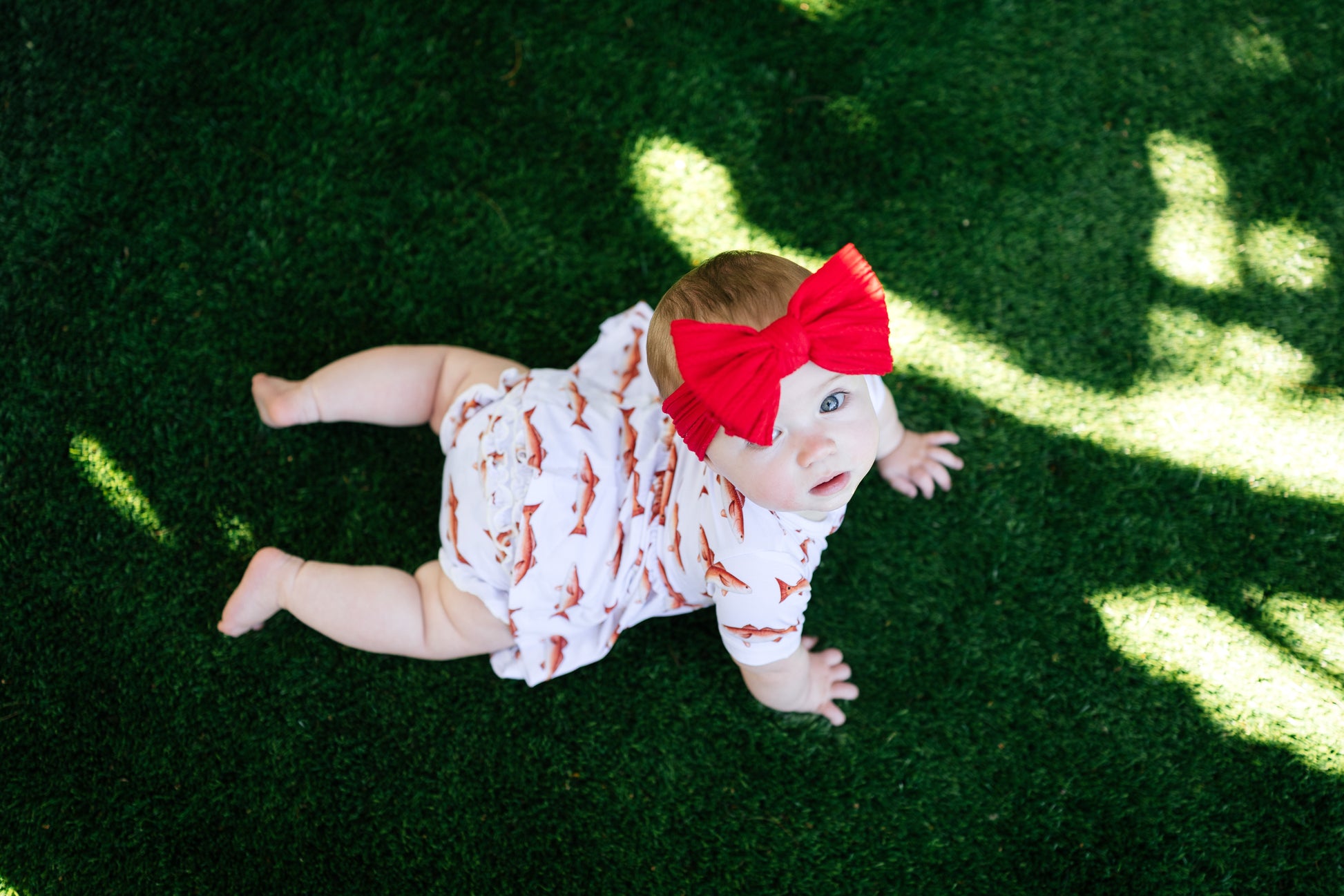 Baby crawling on grass wearing a red bow and white outfit with redfish patterns