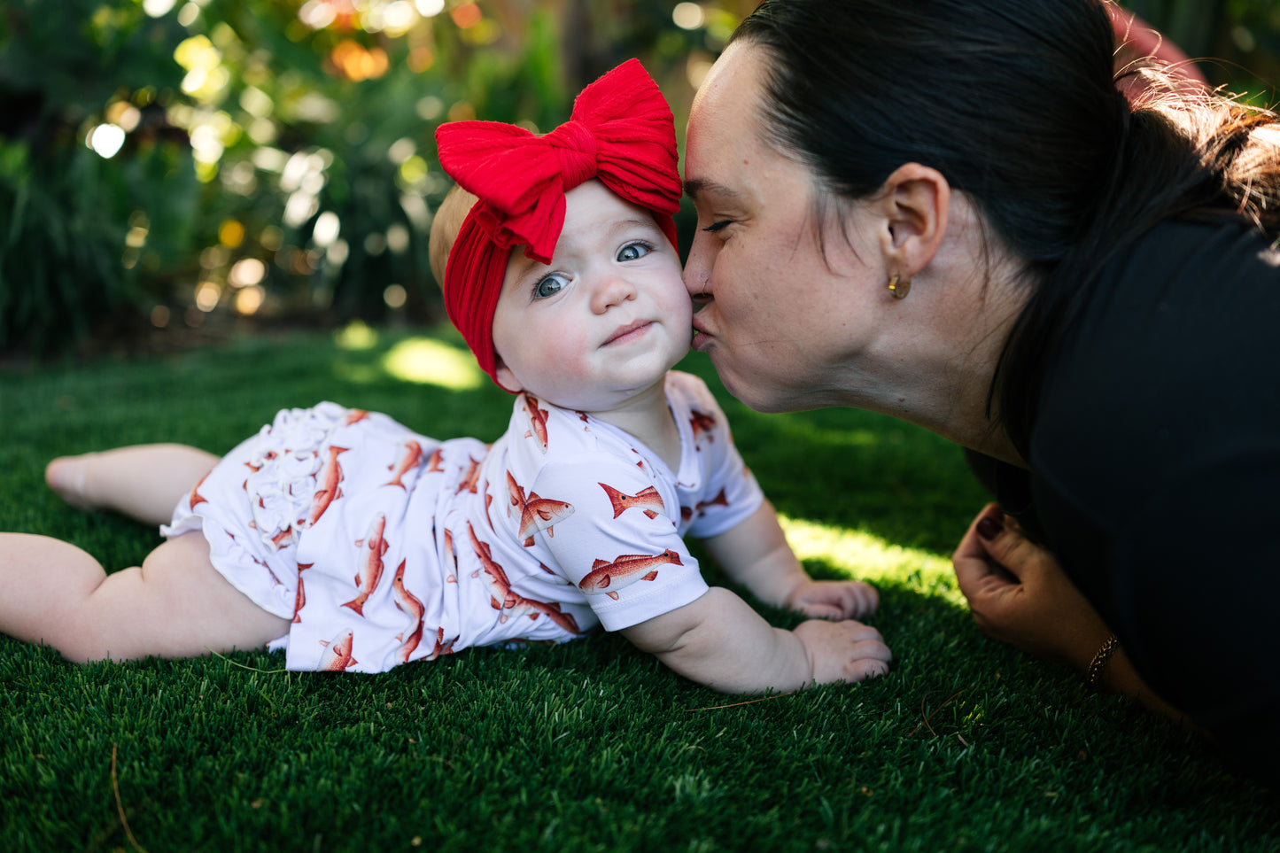 Baby lying on grass wearing a red bow and white outfit, being kissed by her mom