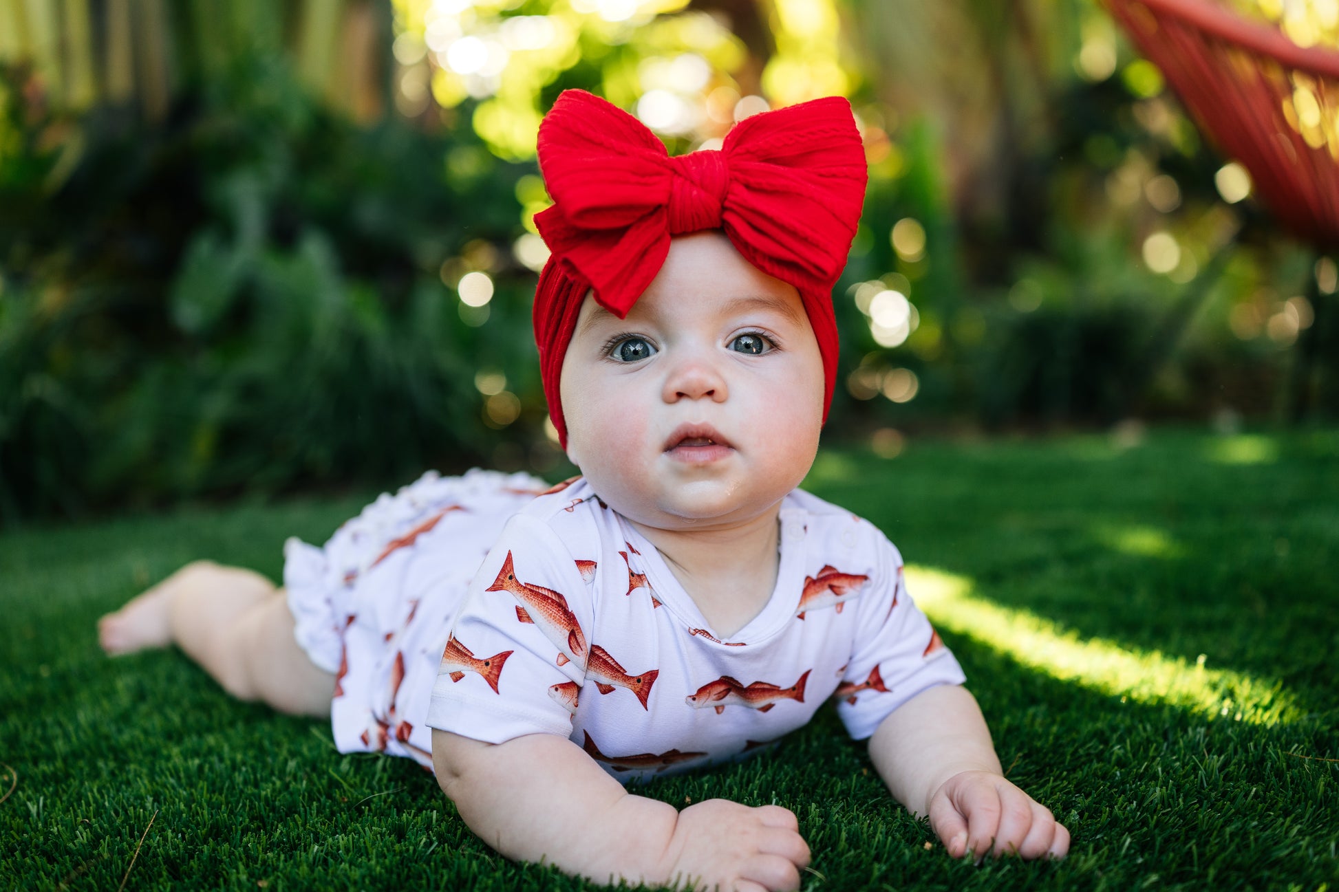 Baby lying on grass wearing a red bow headband and a white outfit with redfish patterns.