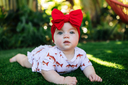 Baby lying on grass wearing a red bow headband and a white outfit with redfish patterns.
