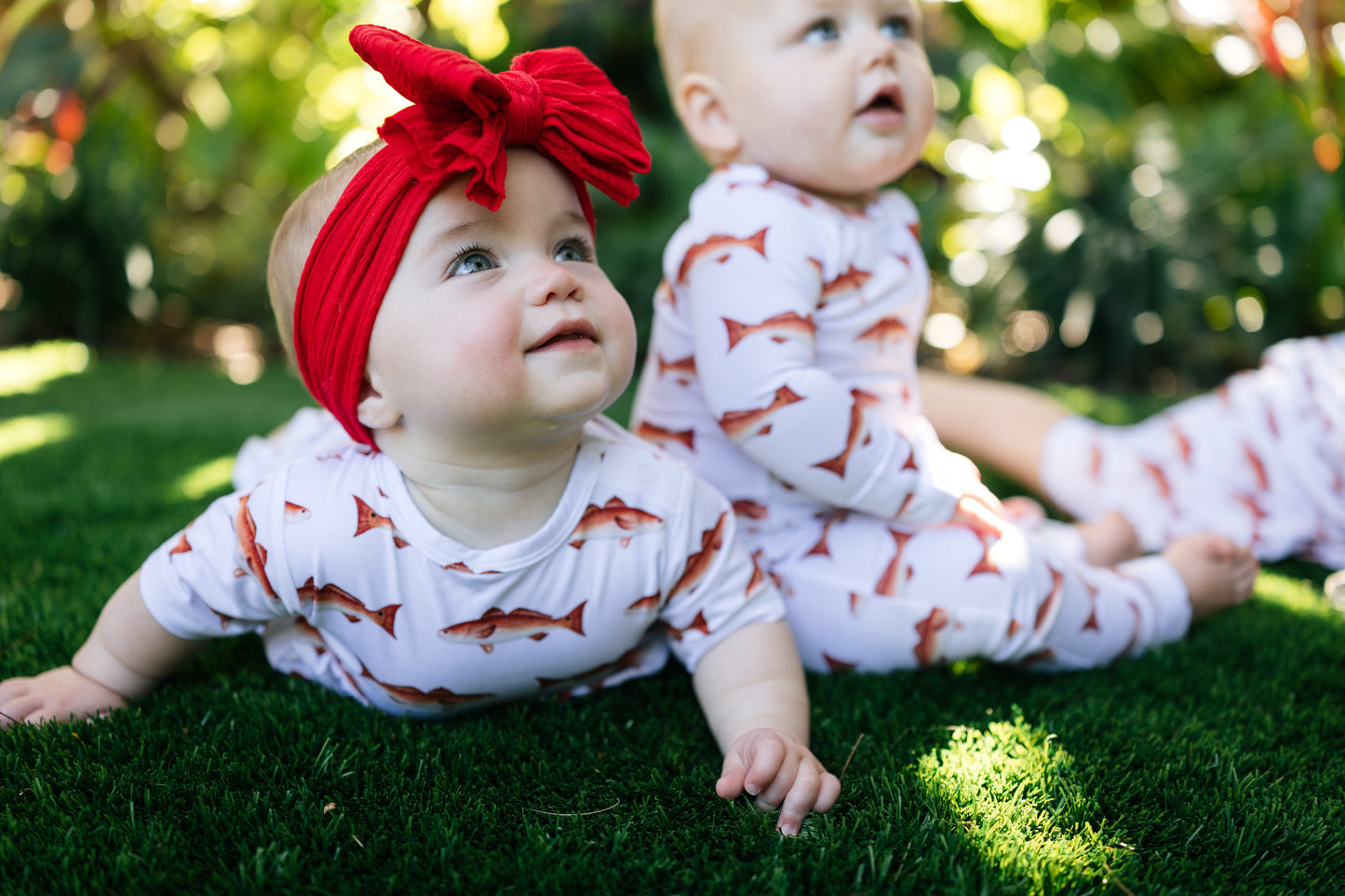 Two babies in matching outfits with a red headband on grass