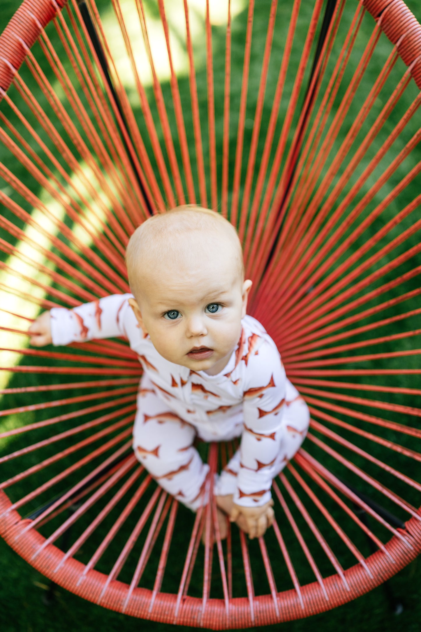 Baby lying on a red woven chair with a blurred green background wearing a redfish zippy