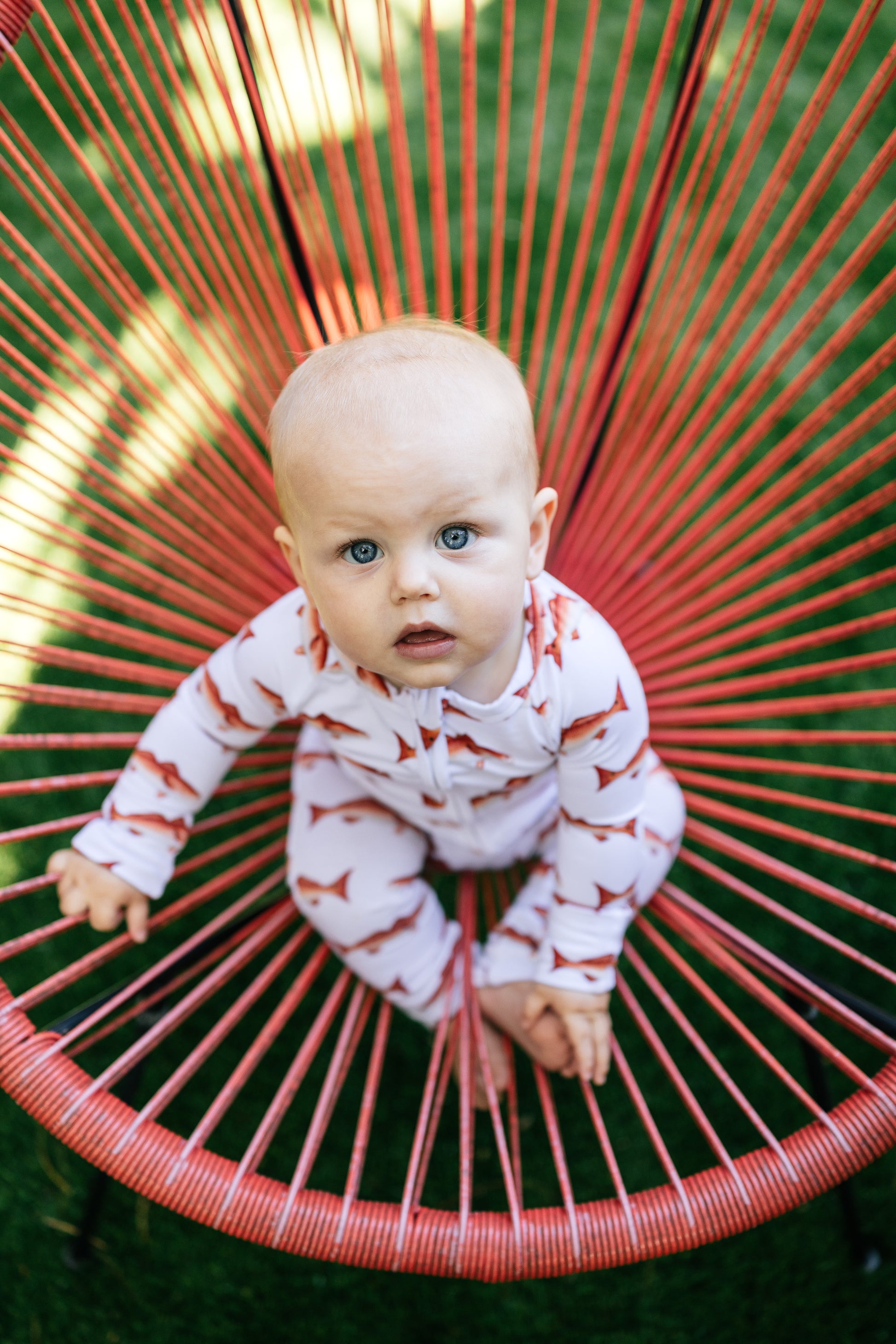 Baby in a redfish zippyying on a red woven chair outdoors.