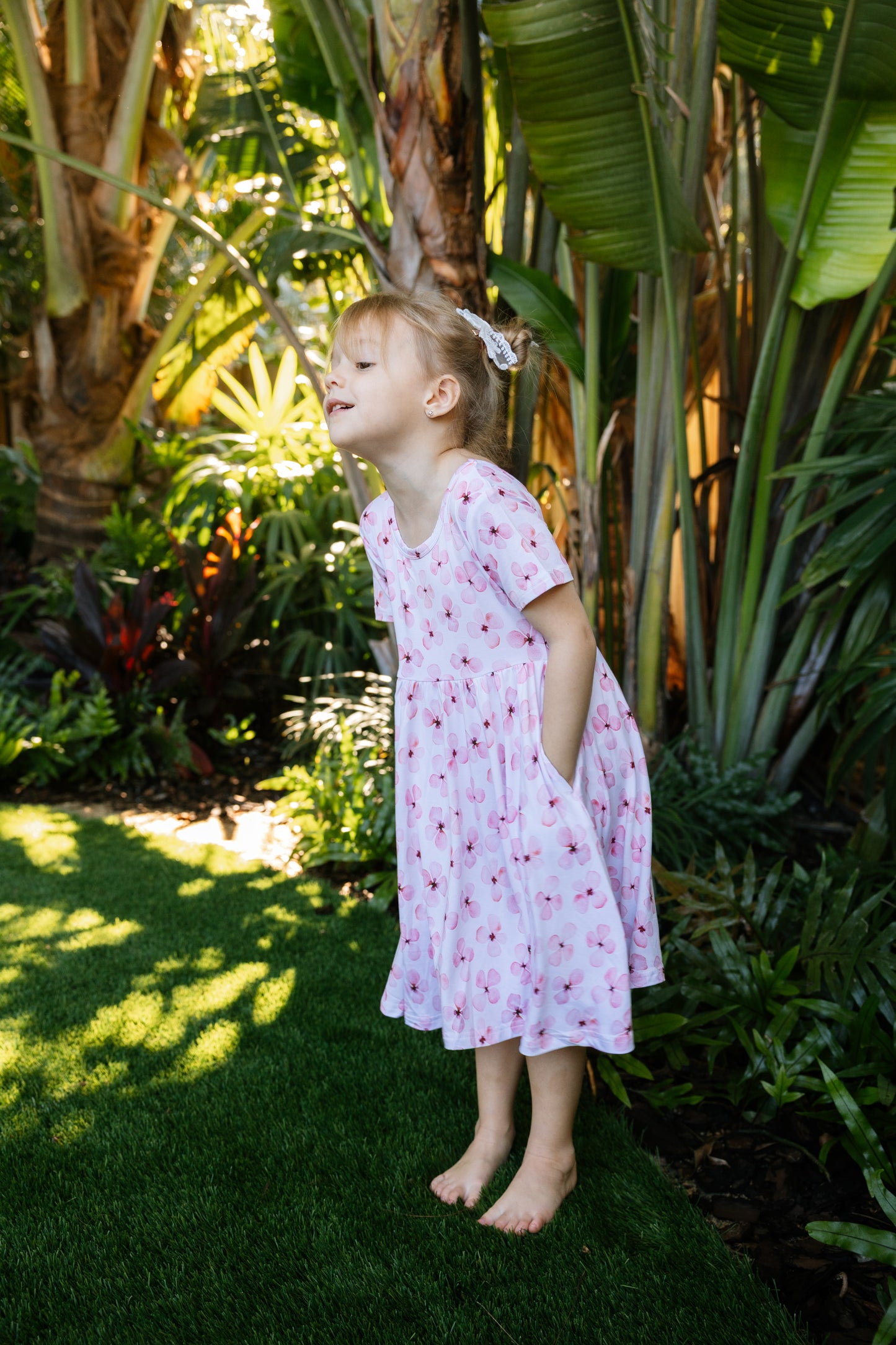 Little girl posing with a tropical background, wearing a white twirl dress with pink flowers short sleeves and pockets