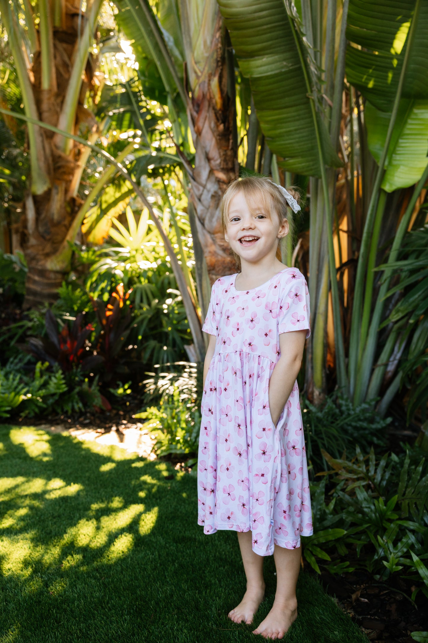 Little girl posing with a tropical background, wearing a white twirl dress with pink flowers short sleeves and pockets