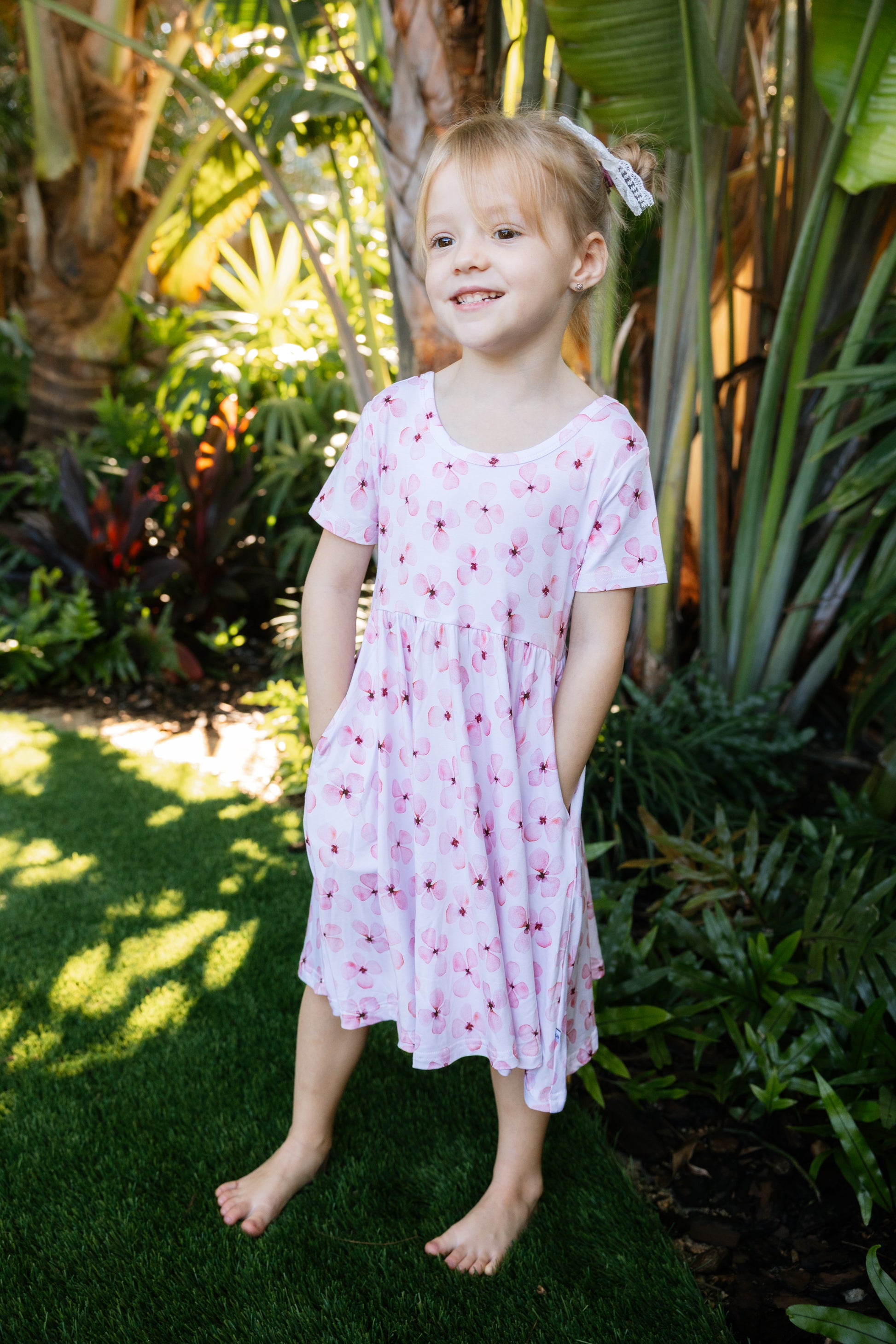 Little girl posing with a tropical background, wearing a white twirl dress with pink flowers short sleeves and pockets