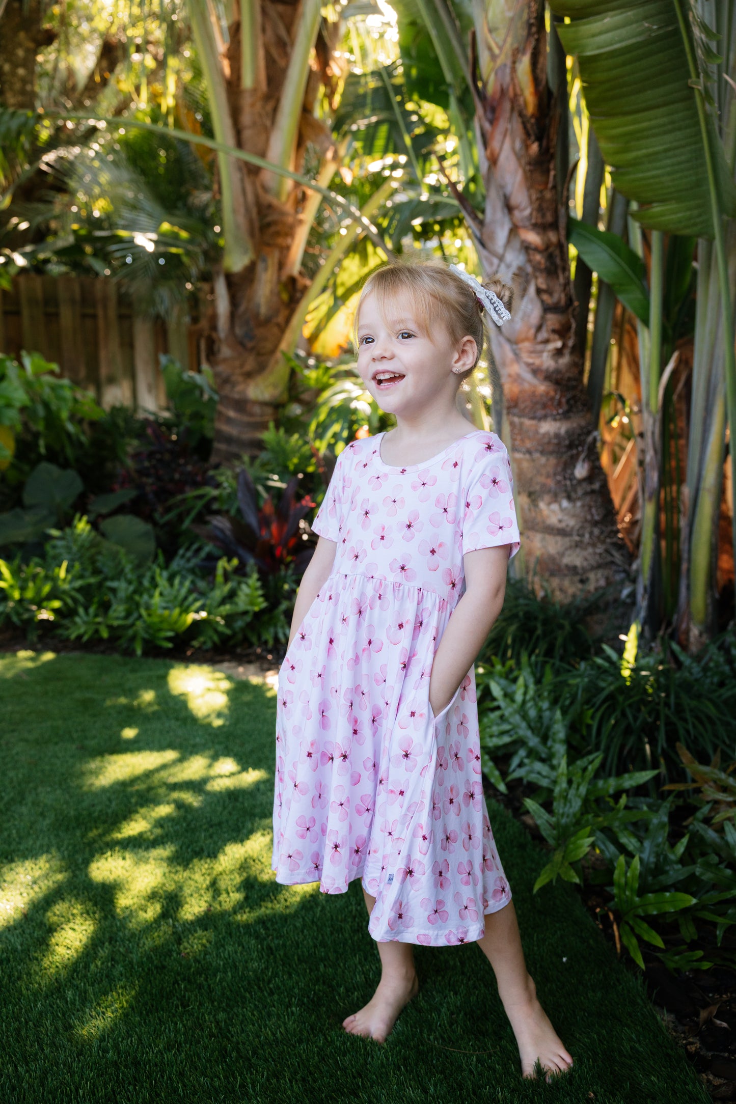 Little girl posing with a tropical background, wearing a white twirl dress with pink flowers short sleeves and pockets
