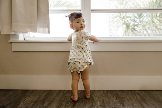 Child standing by a window in a home setting wearing the peplum and ruffle bloomers set with yellow background and permit fish.