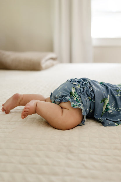 Close-up of infant feet wearing the blue ruffle, peplum and bloomer set with sea turtles laying on a bed