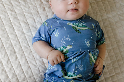 Baby wearing a blue outfit with turtle pattern on a beige textured surface