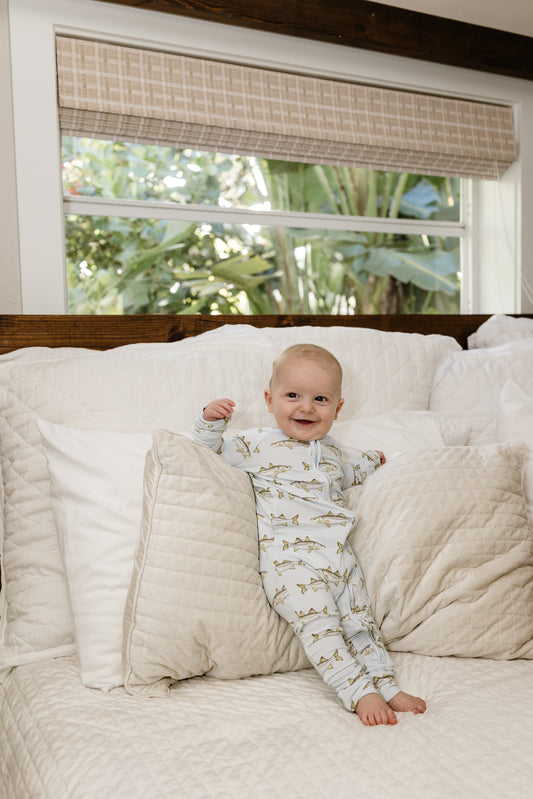 Baby in  snook zippy pajamas sitting on a white bed with a window in the background