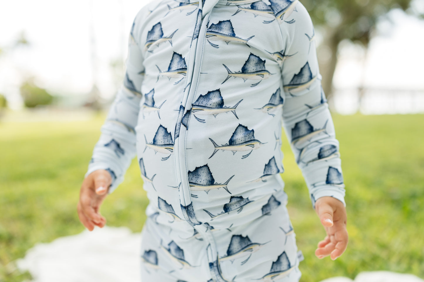 A person outdoors wearing a light blue long-sleeve zip-front top with a repeating sailfish print in dark blue and gray, standing on a grassy lawn.