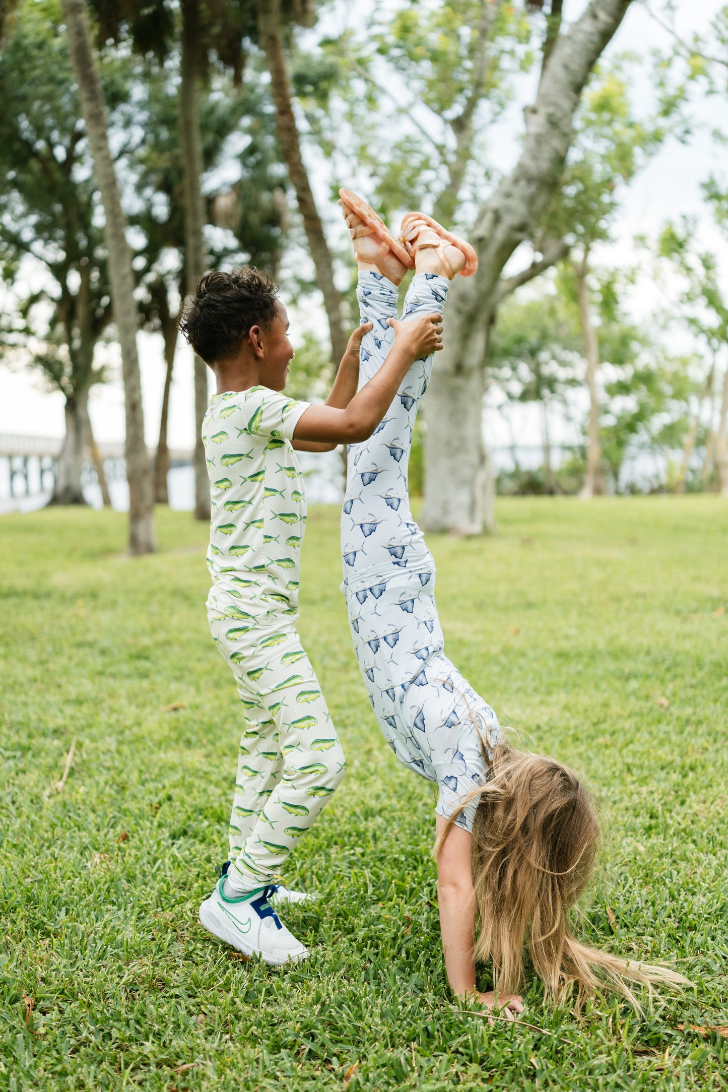 Two children playing outdoors on a grassy park lawn. The boy stands holding up the girl who is doing a handstand, wearing matching light green and white patterned pajamas, while the girl wears light blue and white patterned pajamas.