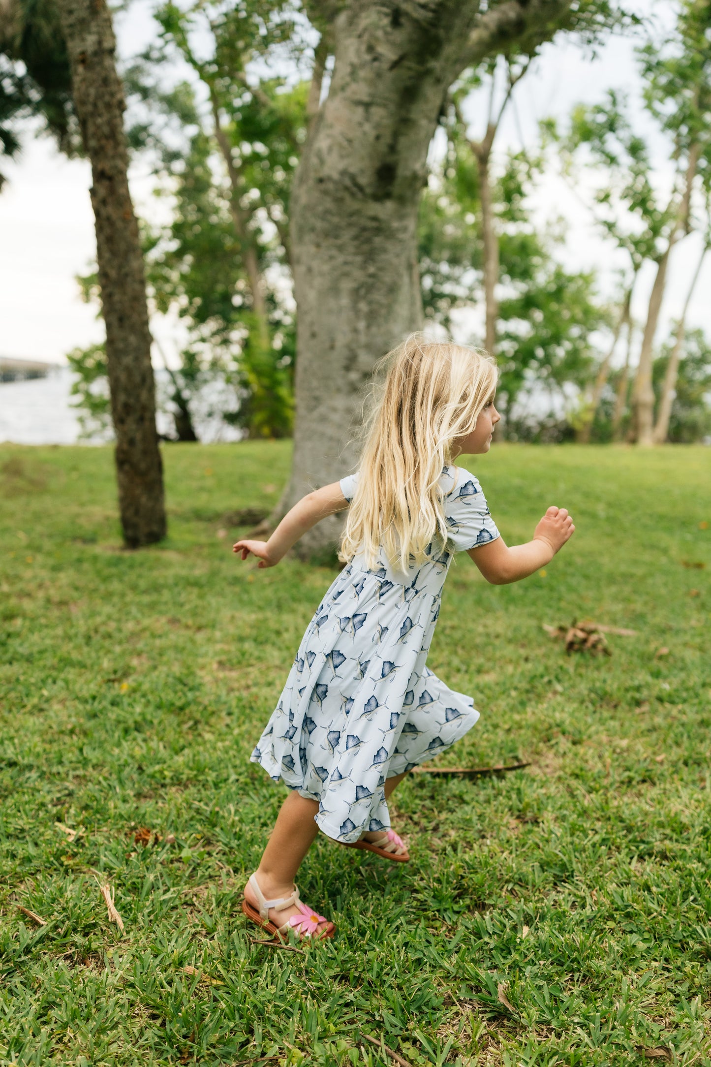 A young blonde girl wearing a white short-sleeve dress with a blue anchor pattern and pink sandals is running on a green grassy lawn in a park with trees.