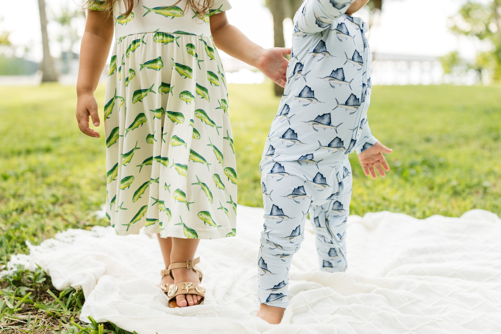 Two children outdoors on a white blanket in a grassy park. One child wears a cream dress with a green fish print, and the other wears a light blue one-piece romper with a dark blue fish print.