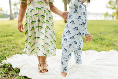 Two children outdoors on a white blanket in a grassy park. One child wears a cream dress with a green fish print, and the other wears a light blue one-piece romper with a dark blue fish print.