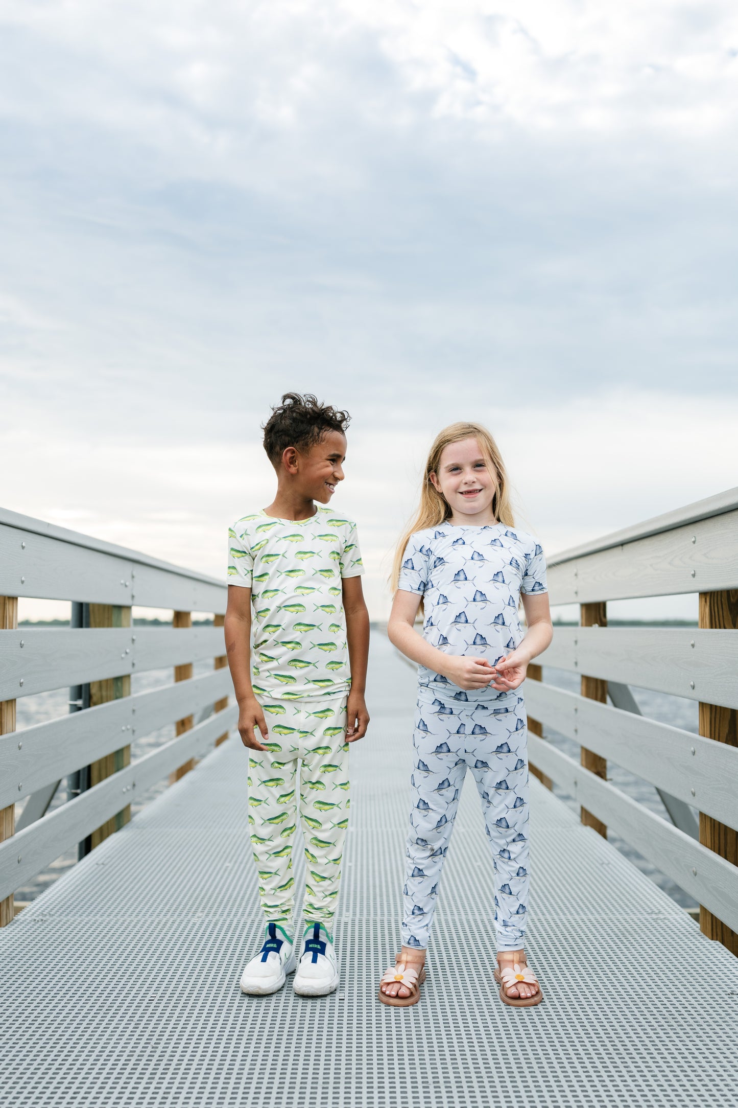 Two children standing on a metal walkway outdoors, wearing matching two-piece pajama sets. The boy's set is white with green dinosaur patterns, and the girl's set is white with blue dinosaur patterns.