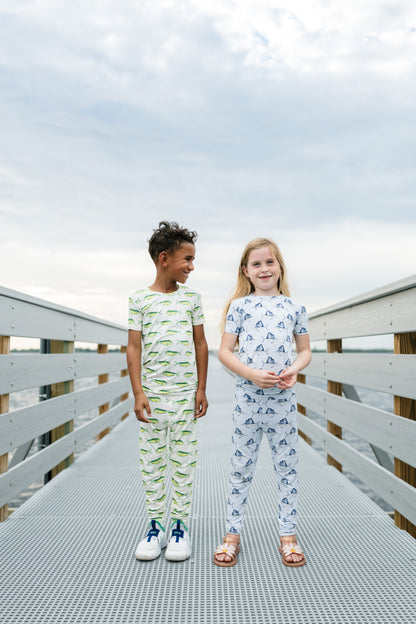 Two children standing on a metal walkway outdoors, wearing matching two-piece pajama sets. The boy's set is white with green dinosaur patterns, and the girl's set is white with blue dinosaur patterns.
