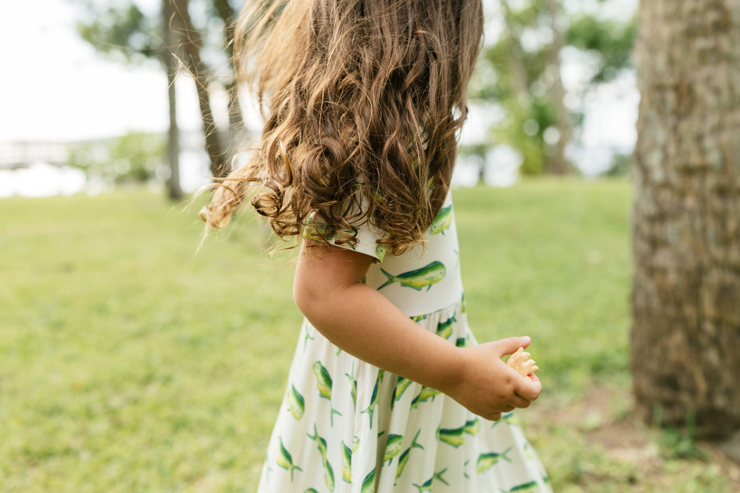 A young child wearing a white dress with a green leaf pattern, standing outdoors in a grassy park next to a tree trunk.