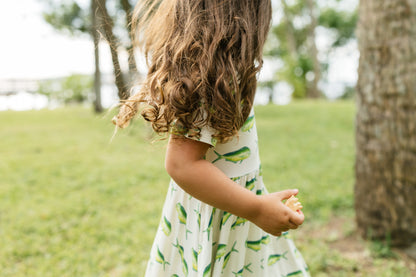 A young child wearing a white dress with a green leaf pattern, standing outdoors in a grassy park next to a tree trunk.