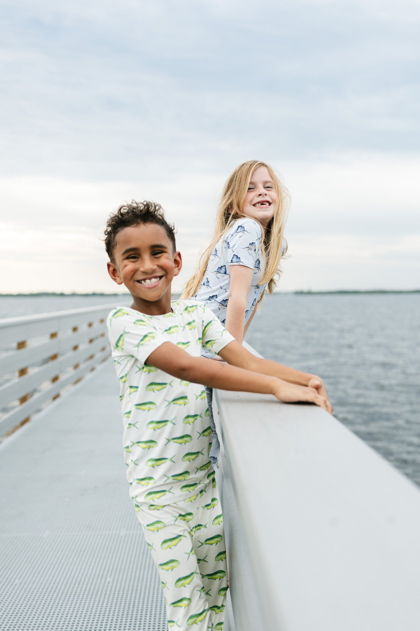 Two children wearing matching printed pajamas stand on a bridge or walkway overlooking water under a cloudy sky.