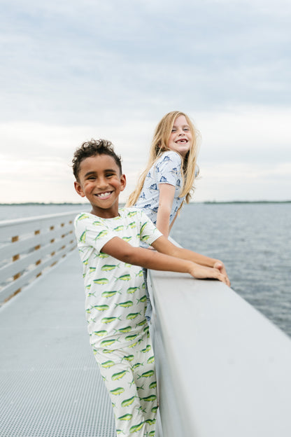 Two children wearing matching printed pajamas stand on a bridge or walkway overlooking water under a cloudy sky.