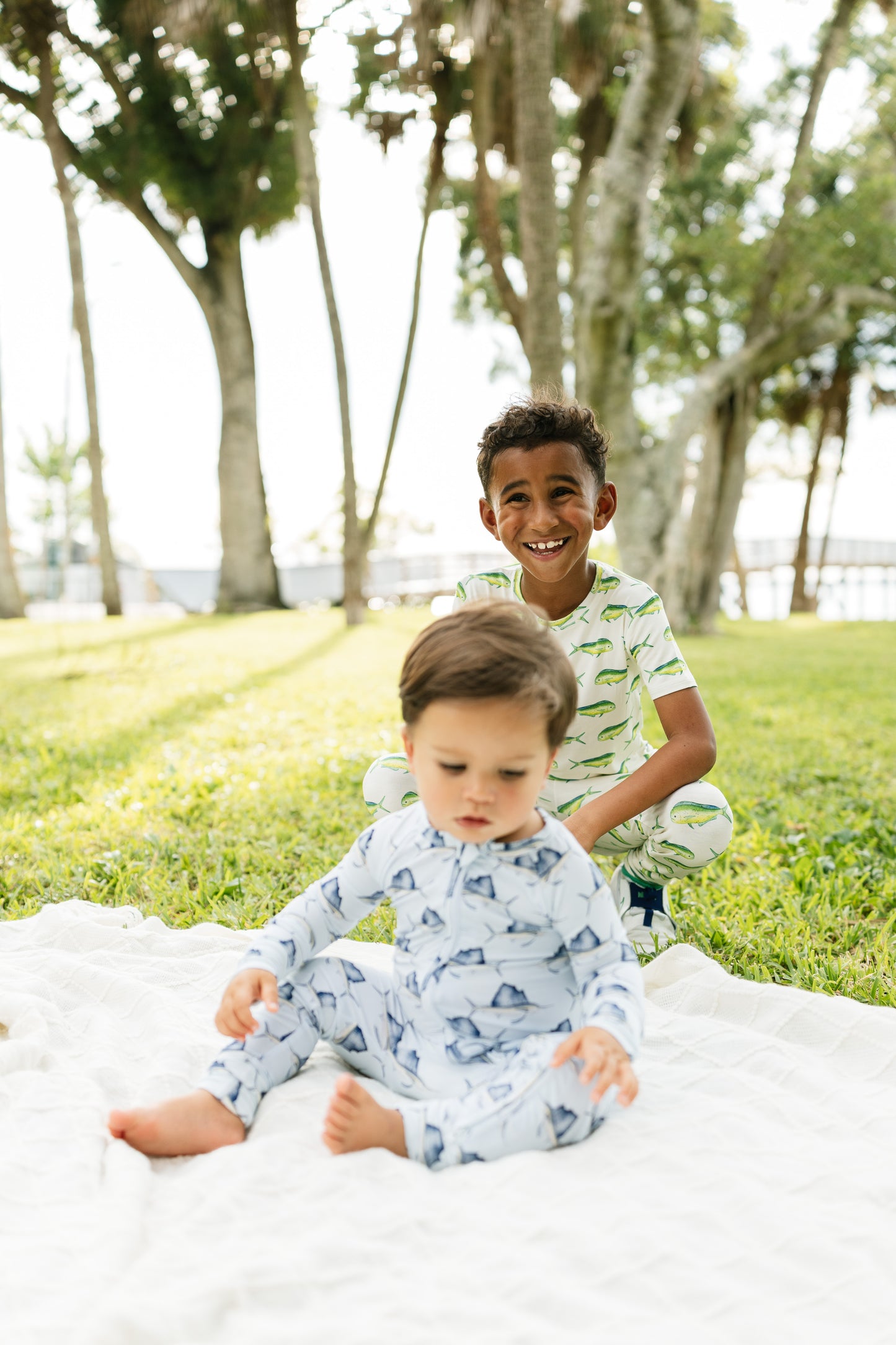 Two children wearing matching patterned pajamas sitting on a white blanket in a park with trees in the background.
