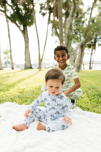 Two children wearing matching patterned pajamas sitting on a white blanket in a park with trees in the background.