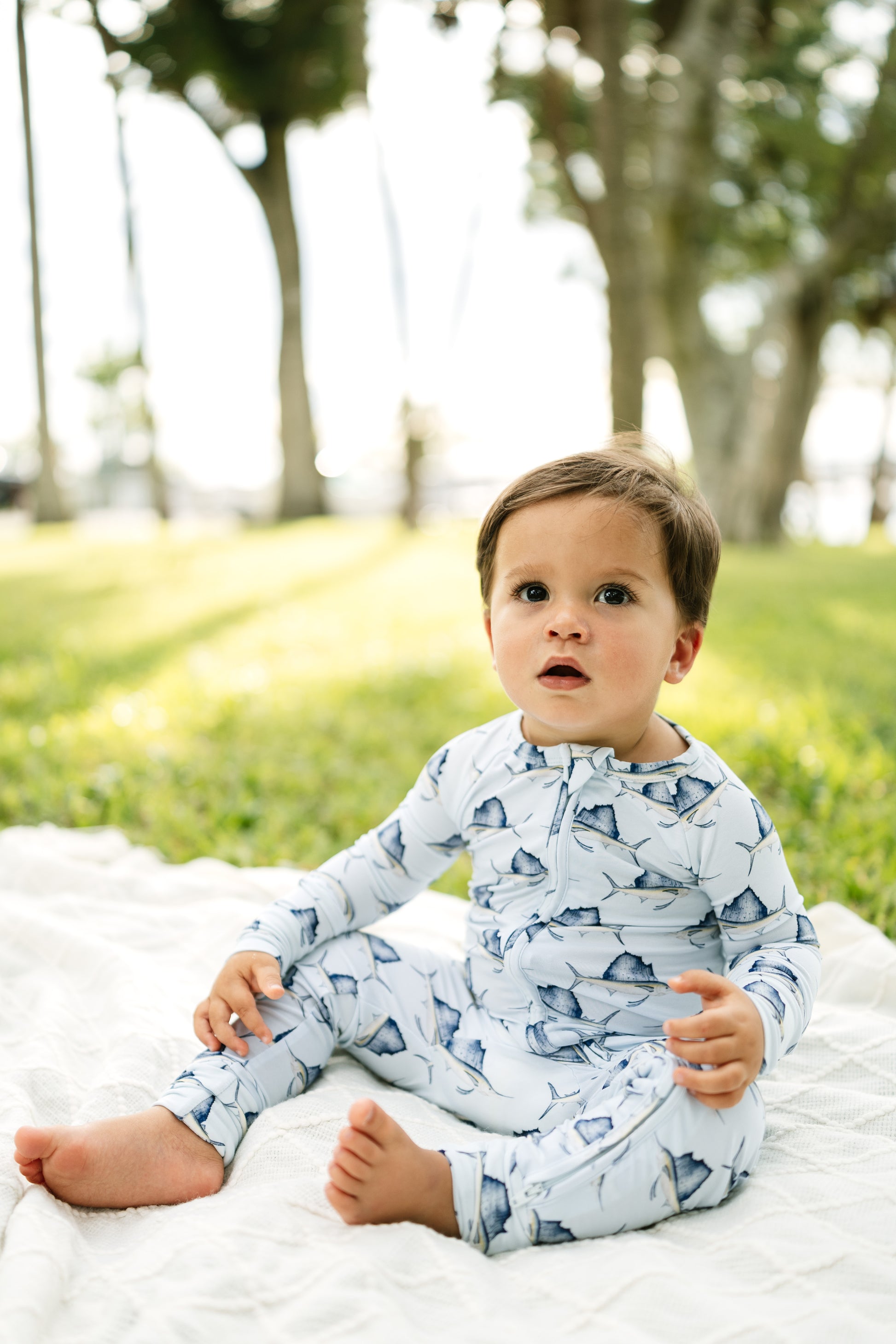 A toddler sitting on a white blanket in a sunny park, wearing a light blue and white long-sleeve one-piece pajama with a shark print.