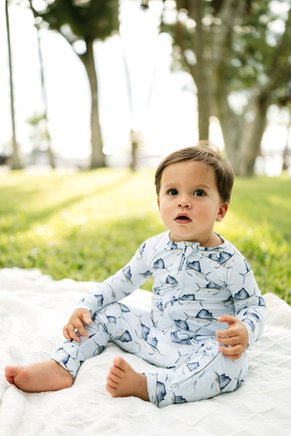 A toddler sitting on a white blanket in a sunny park, wearing a light blue and white long-sleeve one-piece pajama with a shark print.