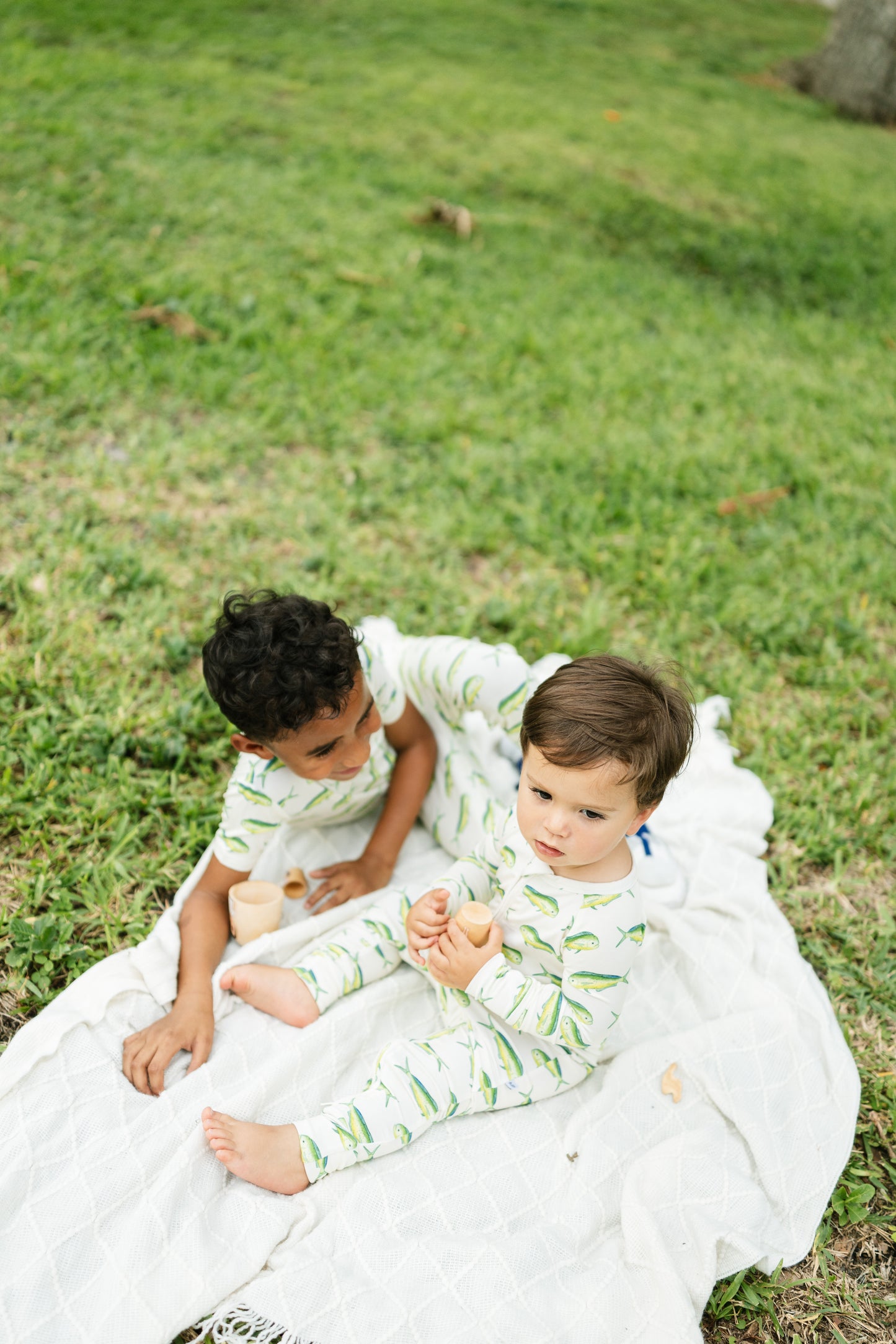 Two toddlers sitting on a white blanket on a grassy lawn, wearing matching light green and white leaf-patterned long-sleeve pajamas.