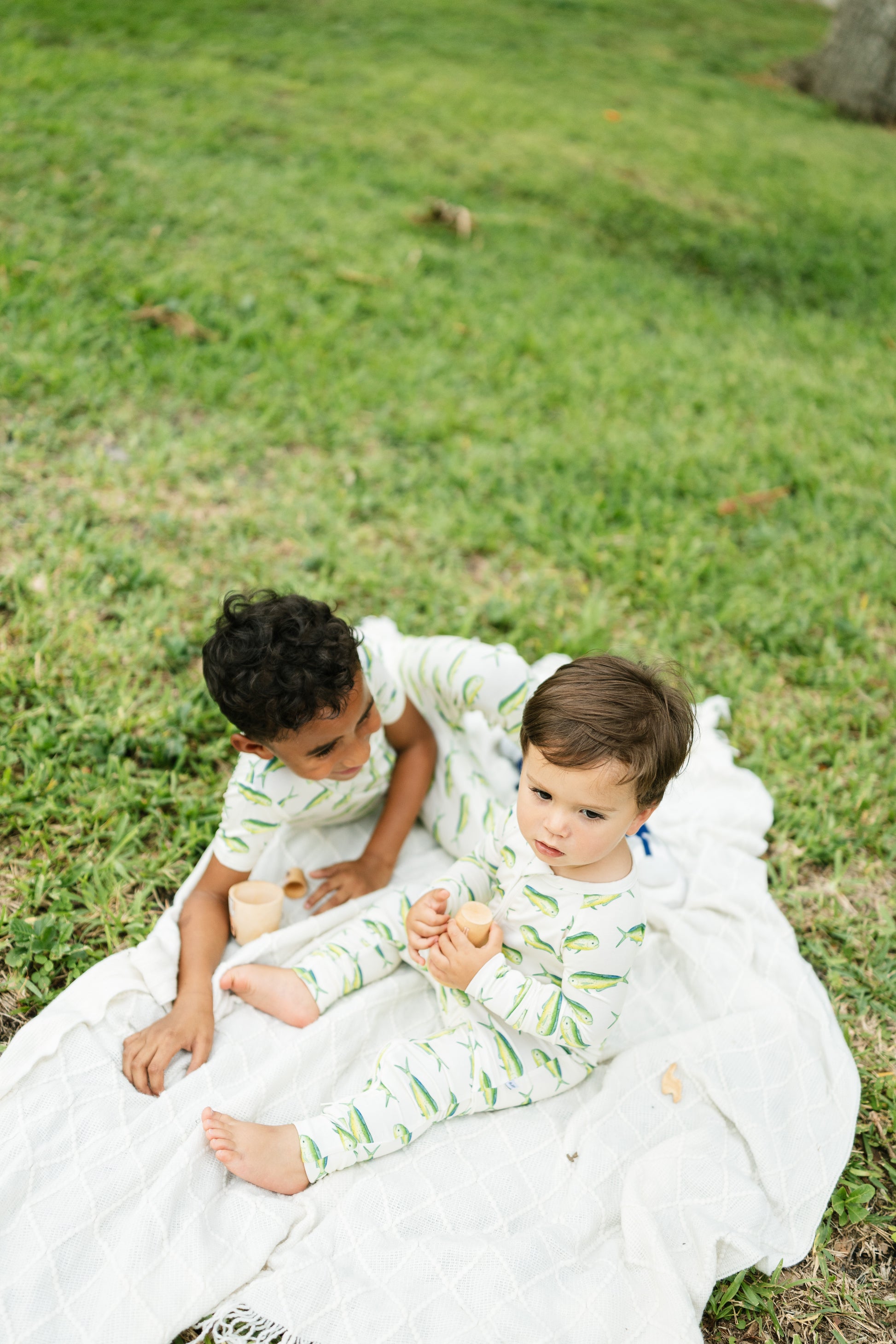 Two toddlers sitting on a white blanket on a grassy lawn, wearing matching light green and white leaf-patterned long-sleeve pajamas.