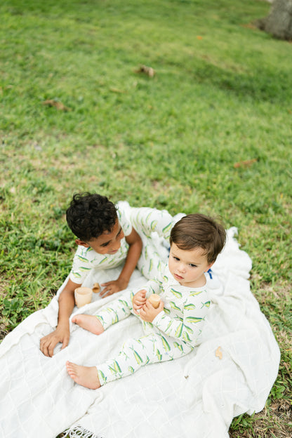 Two toddlers sitting on a white blanket on a grassy lawn, wearing matching light green and white leaf-patterned long-sleeve pajamas.