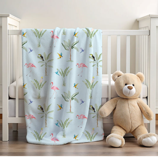 A light blue baby blanket with a tropical print of palm trees, flamingos, and birds, draped over a white crib next to a plush teddy bear.
