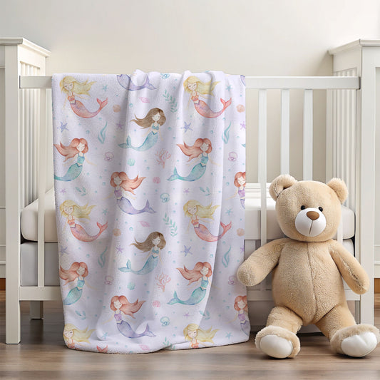 A white baby blanket with a colorful mermaid pattern draped over a white crib, next to a large beige teddy bear toy on a wooden floor.